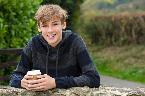 Young happy laughing male boy teenager blond child young adult drinking takeaway coffee outside in fall sunshine