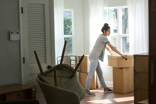 A young mixed race woman, Hispanic and Pacific Islander ethnicity, moving into a house or apartment. She is lifting a cardboard box indoors by a sunny window.
