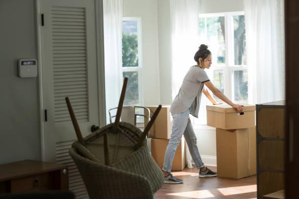 Young mixed race woman moving into new home A young mixed race woman, Hispanic and Pacific Islander ethnicity, moving into a house or apartment. She is lifting a cardboard box indoors by a sunny window. home ownership women stock pictures, royalty-free photos & images