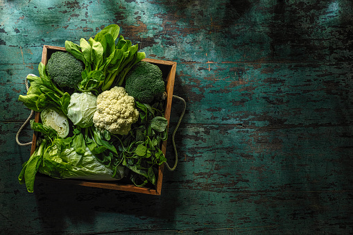 Market fresh green leaf vegetables in an old wooden crate on an old wooden turquoise table. Vegetables include, broccoli, cauliflower, cabbage, spring greens, bok choy and spinach