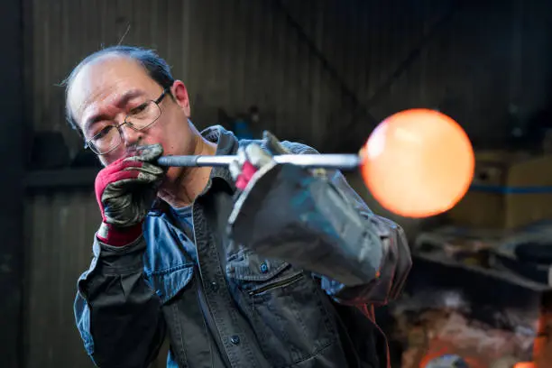 Glassblower shaping molten glass by blowing slowly through a long pipe. Okayama, Japan.