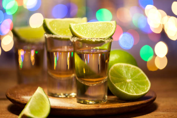 Tequila shots on a bar Gold tequila shots with lime fruits on wooden table on bar lights background tequila shot stock pictures, royalty-free photos & images