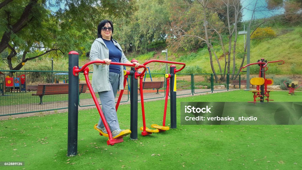Middle aged woman works out an air walker Middle aged woman in gray sport suit tries out on an air walker trainer in her local green park. Happy woman in sunglasses works out at outdoor gym. Promote health and well-being lifestyle concept. Bright sunny morning. Active Lifestyle Stock Photo