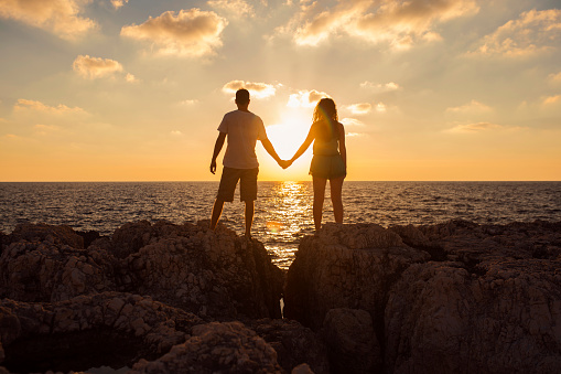 Young heterosexual couple standing on the cliff by the sea, holding hands and enjoying a romantic sunset.