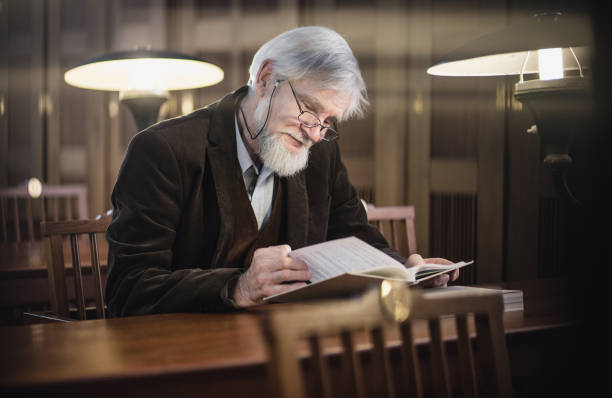 Focused man in eyeglasses reading book at home in morning. Close-up shot of cheerful man reading hardcover book with interest, having rest on weekend. Hobby, education concept