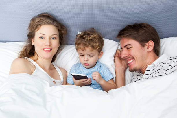 istock Funny boy holding phone lying in bed with parents 643025352