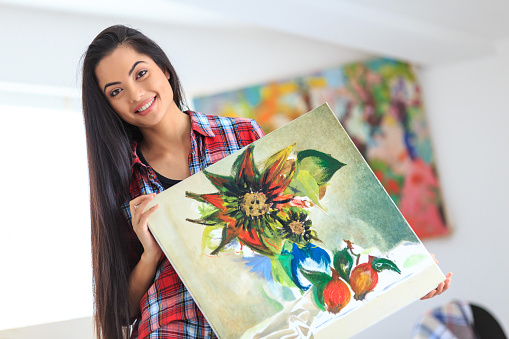 Portrait of beautiful young asian girl holding a painting with flowers in sunny art studio. Wears casual clothes, with long black hair. Looking at camera. Paintings on background.