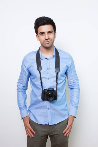 Closeup portrait of content young man looking straight and standing with photo camera hanging on his neck. Isolated front view on white background.