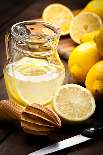 Vertical shot of a jug with infused lemon water and fresh organic lemons on rustic wood table. A wooden citrus squeezer is beside the jug, one lemon is cut in halves. Predominant colors are yellow and brown. Low key DSRL studio photo taken with Canon EOS 5D Mk II and Canon EF 70-200mm f/2.8L IS II USM Telephoto Zoom Lens