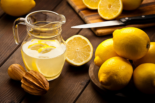 Horizontal shot of a jug with infused lemon water and fresh organic lemons on rustic wood table. A wooden citrus squeezer is beside the jug, one lemon is cut in halves. Predominant colors are yellow and brown. Low key DSRL studio photo taken with Canon EOS 5D Mk II and Canon EF 70-200mm f/2.8L IS II USM Telephoto Zoom Lens