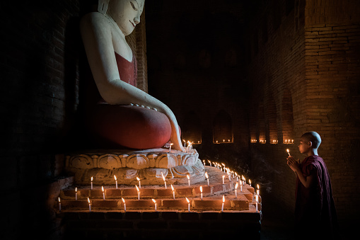 Young burmese buddhist novice monk standing in front of huge buddha statue inside temple surround with burning candles, praying and worshipping. Real People Portrait. Old Bagan, Myanmar, South East Asia.