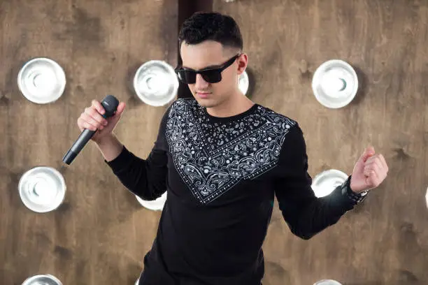 Male singer of rock or pop music dressed in black and sunglasses with microphone performs on scene with lightening projectors on background