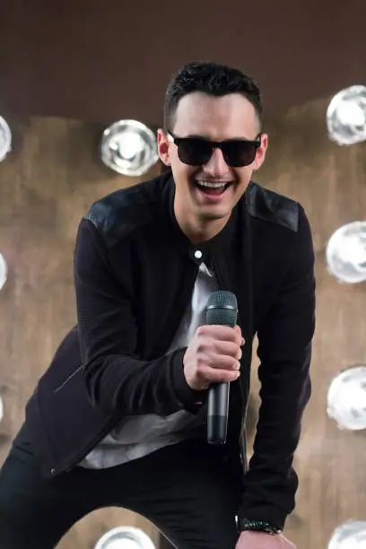 Male singer of rock or pop music dressed in black and sunglasses with microphone performs on scene with lightening projectors on background