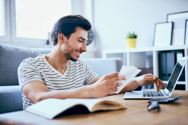 homem feliz, pagando contas em seu laptop na sala de estar - home finances debt tax finance - fotografias e filmes do acervo