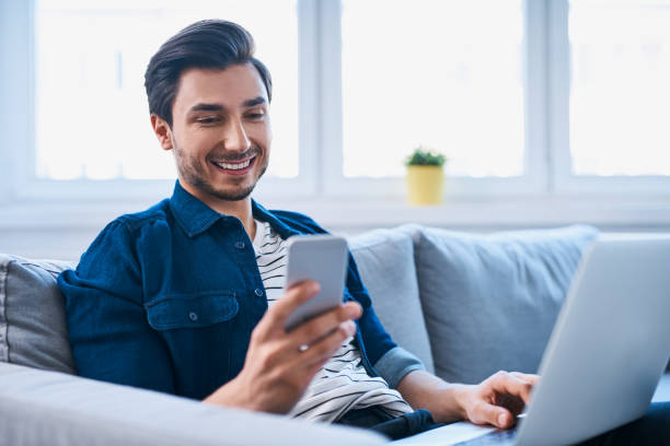 giovane seduto rilassato sul divano con laptop e smartphone - men casual looking at camera one young man only foto e immagini stock