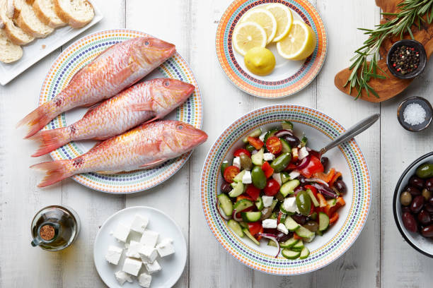 Red mullet fish and mediterranean dishes cooking Raw mullet fish, greek salad and other mediterranean ingredients on white wooden table costus stock pictures, royalty-free photos & images