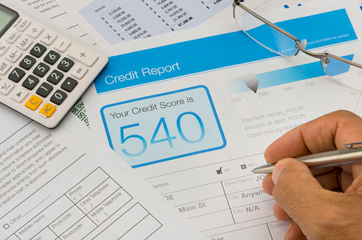 Credit report form on a desk with other paperwork. There are also a pen, glasses and a calculator on the desk. Hand is holding pen