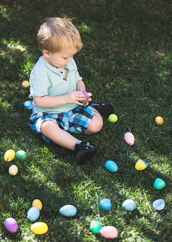 A stock photo of a 2 year old boy sitting on a grass field surrounded by easter eggs. Perfect for designs or articles about easter or easter egg hunts.