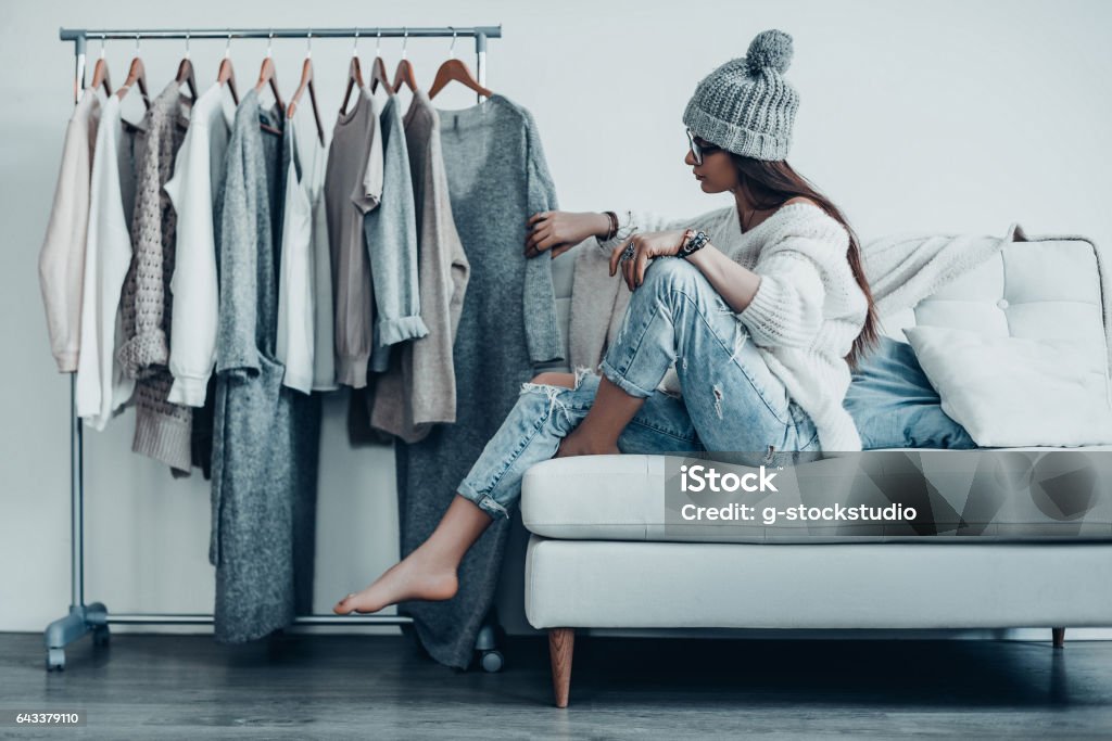 Maybe this one? Thoughtful young woman in casual wear choosing the dress while sitting on the couch at home near her clothes hanging on the racks Closet Stock Photo