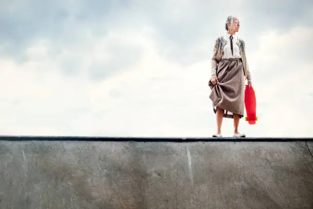 A senior woman attempting to skateboard at a skatepark stands at the edge of a halfpipe debating whether to drop in.   A humorous depiction of being young at heart, exercising with extreme sports even in your 80's.