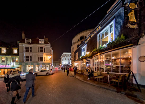 Small groups of people walking in the night time in Brighton, Sussex, UK. Shops, restaurants and bars line the streets. A couple on the left walk away from the camera.
