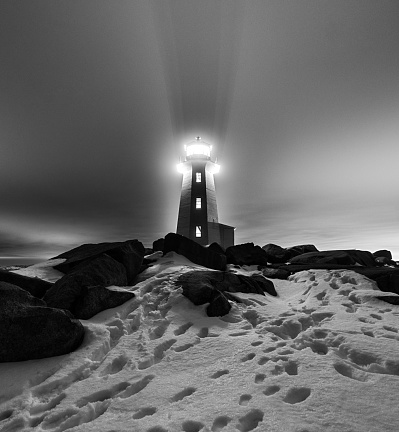 Peggy's Cove Lighthouse is backlit by a bright light, casting a shadow into a thin fog above.  Long exposure shot at high iso.