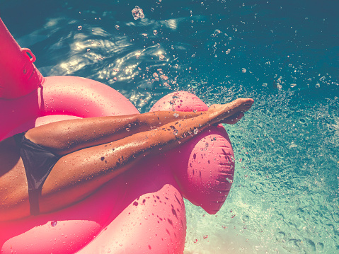 Woman floating on a pink inflatable in swimming pool. Tight crop showing only her legs and feet. She is tanned in turquoise water with water splashing onto her.