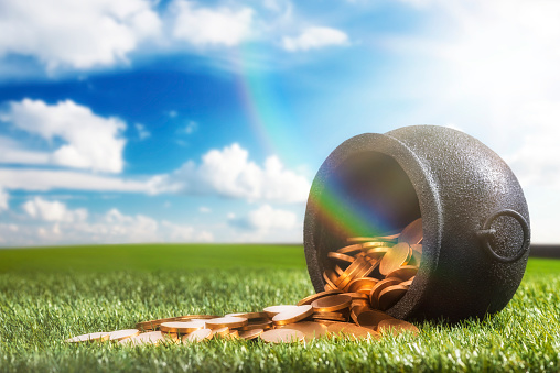 Low angle of a black wrought iron kettle pot that has been knocked over spilling it's gold coins onto the grass, with sun flare and  rainbow. Mythology says it is the secret hiding place of an Irish Leprechaun's pot of gold, some even say he is tricky.