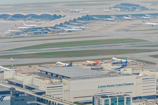 Aerial Hong Kong Airport photo. Photo contains various airplanes from different airlines parked at their gate or taxiing around the airport.
