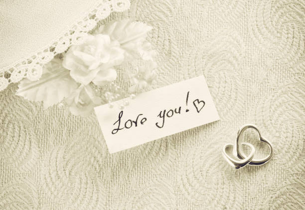 wedding invitation, valentine day concept, sepia toned card with a clothes detail lace, boutonniere, two silver heart pendant, a note with confession of love