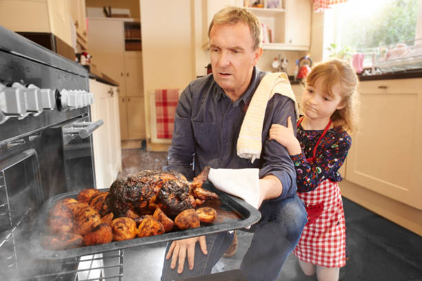 roast dinner fail a mature man pulls the chicken or turkey out of the oven only to find it burnt and ruined. His little daughter consoles him. burnt stock pictures, royalty-free photos & images