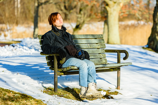 Young adult female dressed in black and blue, basking in sunshine on park bench in winter or early spring. Snow on the ground. Sunny warm day with some thaw around the tree and bench. Camera in lap.