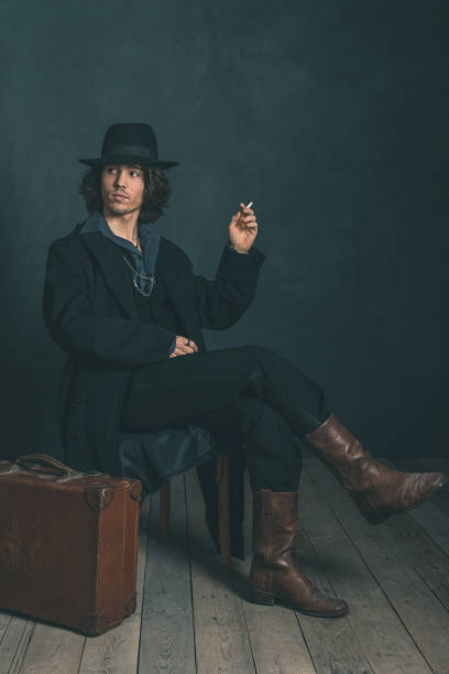 istock Artistic alternative vintage man wearing black hat and coat. Smoking cigarette. Sitting on wooden chair in studio. 643625842