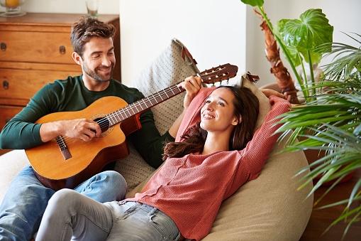 Shot of a young man playing guitar for his girlfriend while relaxing in their living room