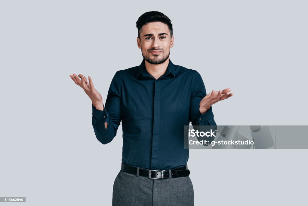 Mystery! Confused young man shrugging shoulders and making face while standing against grey background Confusion Stock Photo