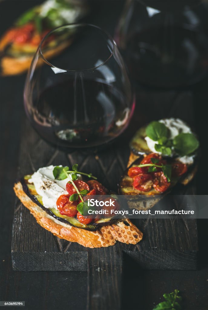 Brushetta with eggplant, tomatoes, garlic, cream-cheese, arugula, glass of wine Wine and snack set. Brushetta with roasted eggplant, tomatoes, garlic, cream cheese, arugula and glass of red wine on wooden board over dark background, selective focus. Slow food, party food concept Bruschetta Stock Photo