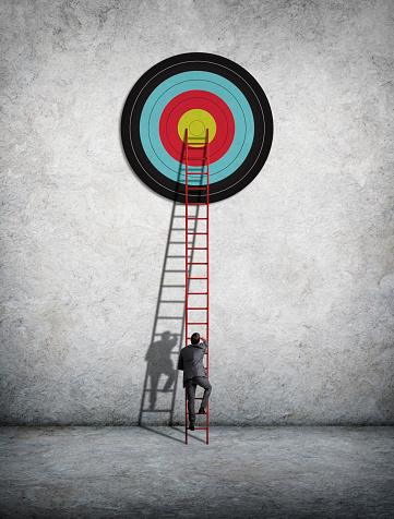 A businessman climbs a tall red ladder as he attempts to reach a large target above him. A strong shadow of the businessman and the ladder is cast on to the wall.