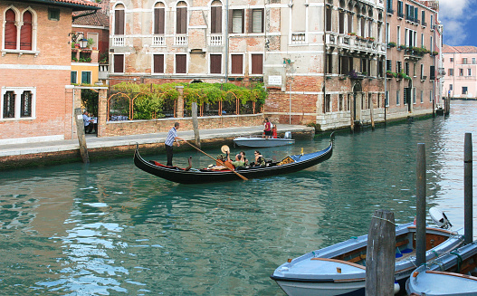 Tourists taking a gondola tour and eating in outdoor cafe in Venice, Italy. Rowing gondolier with an oar, aquamarine canal water, moored motorboats, historic red brick buidings, green trees and blue sky with clouds are in the image.