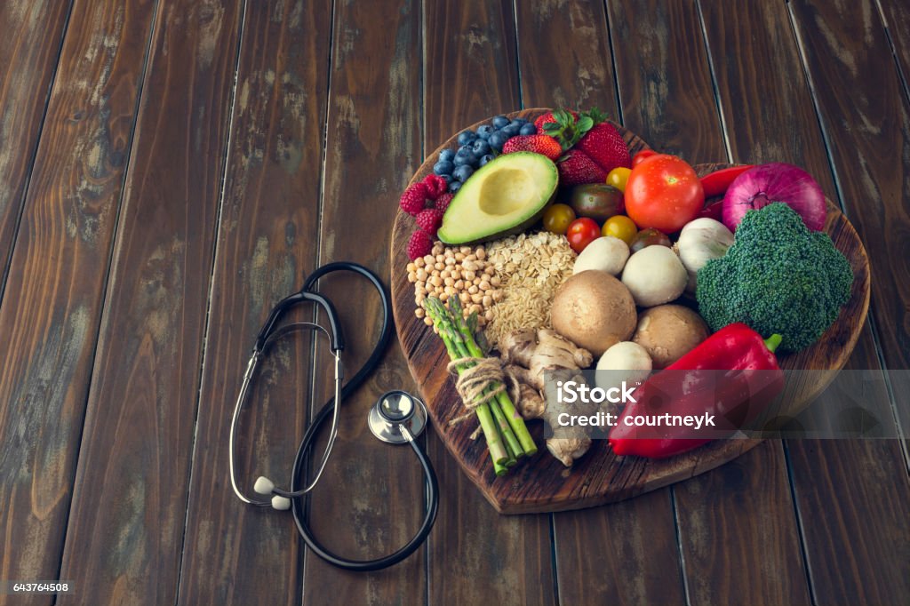 Healthy food on a heart shape cutting board. Healthy food on a heart shape cutting board. Love of food concept with fruit, vegetables, grains and high fibre foods. Rustic wood textures. There is also a stethoscope Heart Shape Stock Photo