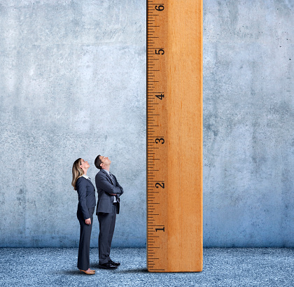A businesswoman and businessman stand at a profile to the camera as they looks up at a large wooden ruler that is standing on its end inside a large room.