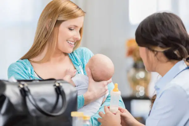Female lactation consultant talks with new mom. The mother is holding her baby girl. The consultant is holding a baby bottle.