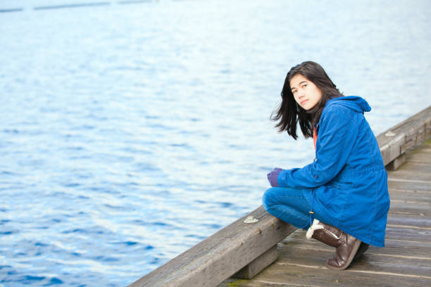 Sad, lonely biracial  teen girl on wooden pier by water Biracial teen girl in blue jacket and boots with lonely or sad expression crouching down on wooden pier over water's edge, looking at camera sad girl crouching stock pictures, royalty-free photos & images