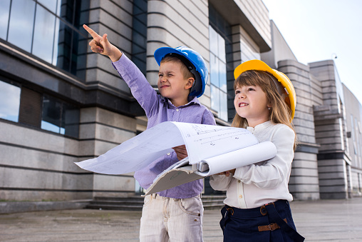 Small architects standing in front of a building and planning a project while boy is pointing up. Focus is on girl.