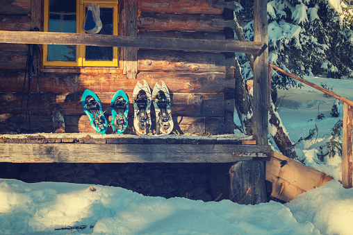 Snowshoes are on the porch of an old log cabin in the warm light of the evening sun. Winter adventure in the wild.
