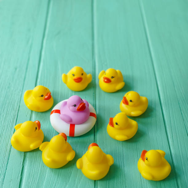 istock Group of yellow rubber ducks gathering around a different purple duck that is sitting on a life saver rubber ring, on a turquoise coloured wooden background. 643863378