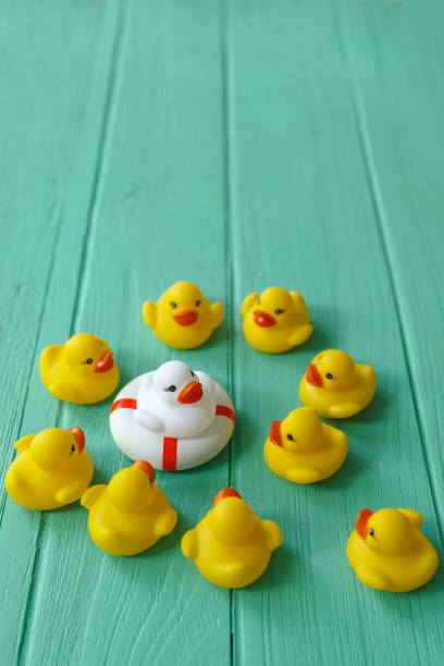 istock Group of yellow rubber ducks gathering around a different white duck that is sitting on a life saver rubber ring, on a turquoise coloured wooden background. 643863390