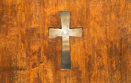 Cross shape hole in a church wooden fence with sun and sky on the background