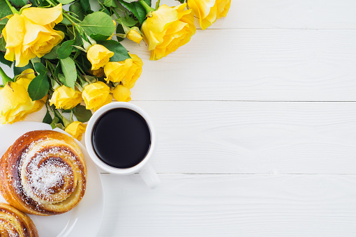 Cup of coffee with bouquet of yellow roses, flat lay, top view. Two spiral tasty buns with coconut chips, cup of black coffee and bouquet of yellow roses. Delicious breakfast, sweet rolls and coffee.