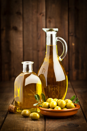 Vertical shot of two olive oil bottles of different shapes and a brown bowl filled with green olives on rustic wood table. Predominant colors are gold, green and brown. DSRL studio photo taken with Canon EOS 5D Mk II and Canon EF 70-200mm f/2.8L IS II USM Telephoto Zoom Lens