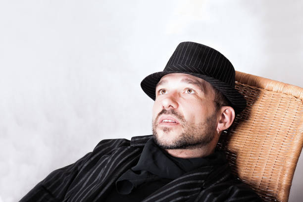 Portrait of handsome bearded man with pierced ear in black hat and cravat sitting in a chair.